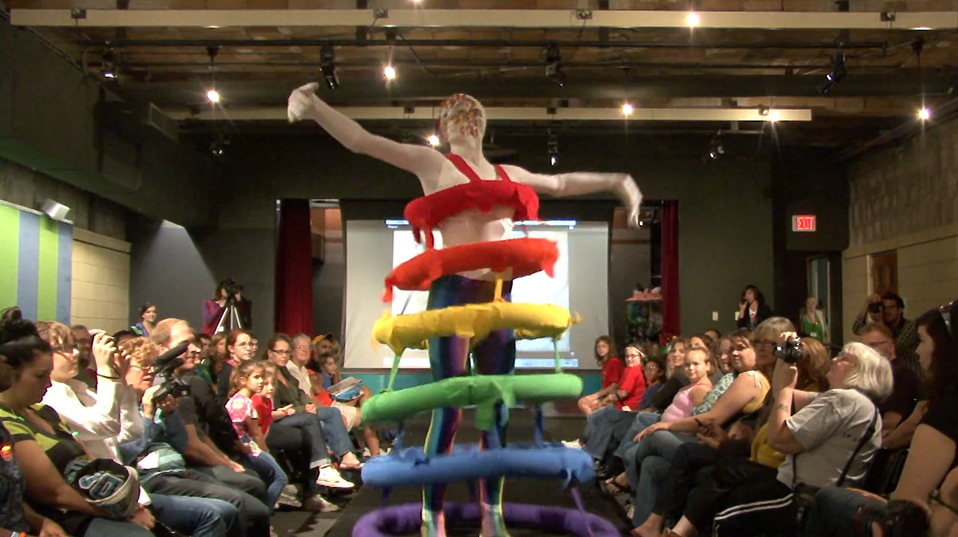 Performance art with a person in a colorful, tiered costume on a stage, surrounded by an audience.